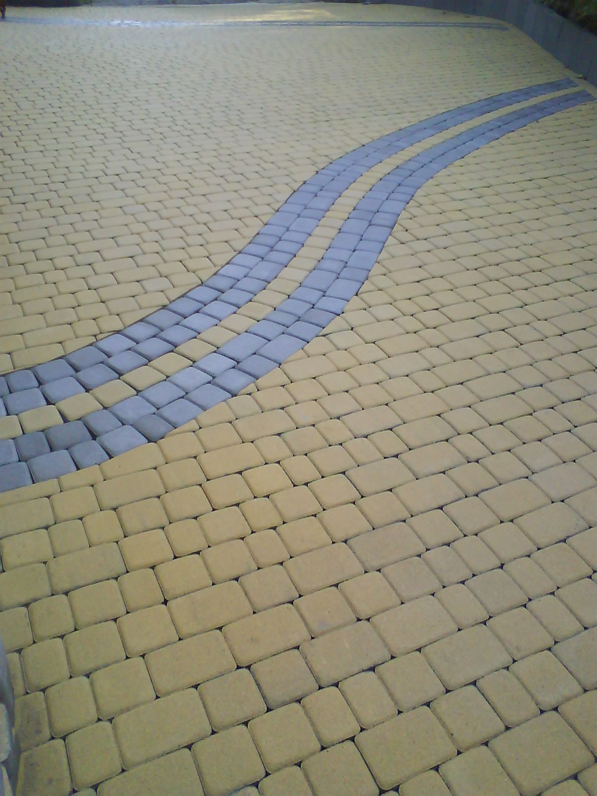 Ułożona nawierzchnia z kostki brukowej w odcieniach żółci i szarości, tworząca wzór dwóch równoległych, falujących pasów.